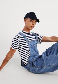 Salopette en denim avec bretelles ajustables, portée par-dessus un t-shirt à rayures bleu marine et blanches. Casquette noire et tatouage visible sur le bras. Look décontracté et ample.