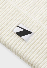 Un bonnet en maille crème avec une texture côtelée, présentant une étiquette de marque noire et blanche sur le côté. Design simple et fonctionnel.