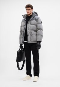 Chaqueta acolchada gris con capucha, que presenta un cierre de botones y dos bolsillos frontales. Lleva puesta sobre un suéter negro, pantalones negros y zapatillas blancas.
