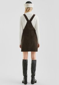Frau mit einem weißen Kabelstrickpullover, einem dunkelbraunen Cord-Pinafore-Kleid, schwarzen kniehohen Stiefeln und einem schwarzen Hut mit weißem Fellrand, Rückansicht.