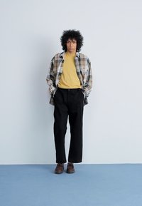 Geruite blouse met lange mouwen over een gele t-shirt, gecombineerd met zwarte wijdvallende broek en bruine schoenen. Neutrale achtergrond met een blauwe vloer.