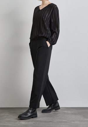 Personne portant un chemisier noir à fines rayures, manches longues, un pantalon noir droit et des bottines en cuir noir, debout contre un mur gris.