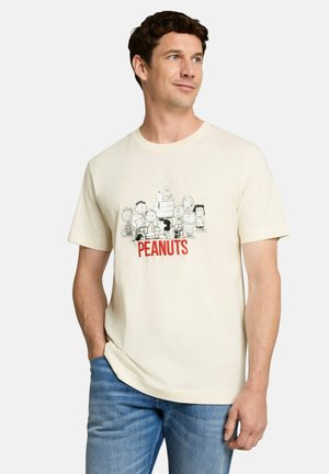 Mann trägt cremefarbenes T-Shirt mit schwarz-weißen Peanuts-Charakteren und rotem "PEANUTS" Text, kombiniert mit blauen Jeans, sieht nach rechts mit der Hand in der Tasche.