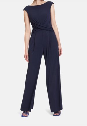 Vrouw draagt een marineblauwe mouwloze jumpsuit met een gedraaide taille en wijde pijpen, gecombineerd met blauwe sandalen met bandjes.