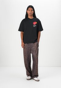 Svart oversized t-skjorte med logo i rødt og oransje på brystet, kombinert med brune bukser med leopardmønster og hvite stripes på sidene.