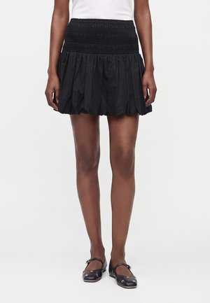 Femme portant une mini-jupe noire froncée avec une ceinture smockée, associée à des ballerines Mary Jane noires cloutées et un haut blanc.