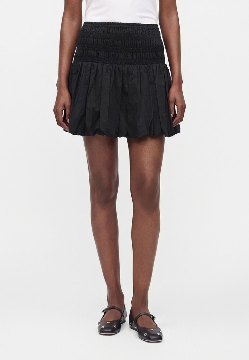 Femme portant une mini-jupe noire froncée avec une ceinture smockée, associée à des ballerines Mary Jane noires cloutées et un haut blanc.