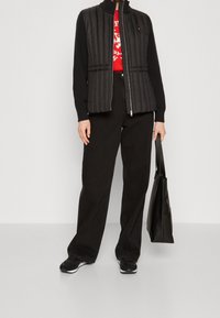 Gilet trapuntato nero con colletto alto, abbinato a una maglietta grafica rossa e pantaloni neri a gamba larga. Gli accessori includono una borsa a tracolla nera e sneakers.