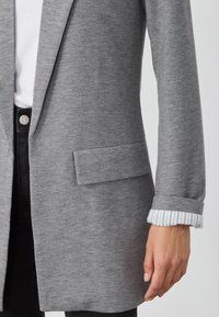 Blazer grigio con una texture liscia, dotato di colletto a sovrapposizione, taschino singolo sul davanti e dettaglio a righe nei polsini.