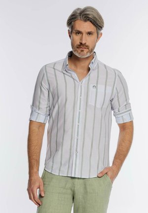 Mężczyzna z siwymi włosami i brodą, ubrany w białą koszulę zapinaną na guziki z pionowymi paskami i podwiniętymi rękawami oraz jasne zielone spodnie, stoi z jedną ręką w kieszeni.