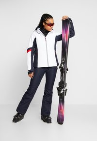 Ski outfit bestående af en hvid og marineblå jakke med røde accenter, sammen med marineblå bukser og sorte skistøvler, der holder farverige ski.