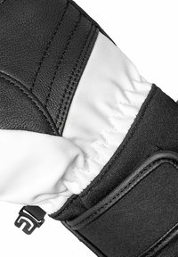 Schwarze Leder- und weiße Stoffhandschuhe mit texturierten Nähten, verstellbarem Riemen und gerafftem Stoff am Handgelenk. Sichtbares Detail der robusten Konstruktion.