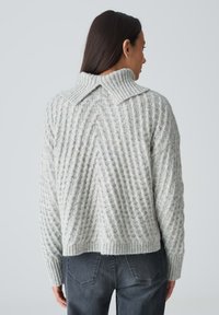 Grau gestrickter Pullover mit einem strukturierten Rautenmuster, geripptem Kragen und Bündchen. Verkürzte Länge, getragen mit hochgeschnittenen grauen Jeans.