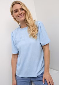 Top corto azzurro chiaro, testurizzato, con scollatura arricciata; forma rotonda, vestibilità comoda, abbinato a jeans in denim chiaro.