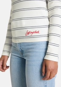 Langärmliges Hemd in Off-White mit marineblauen Streifen, ausgestattet mit einem gestickten roten Logo am Saum. Kombiniert mit hellblauen Jeans.