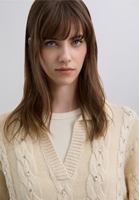 Pull crème en maille torsadée avec col en V, orné de strass décoratifs et d'un motif texturé, porté par-dessus un haut côtelé beige clair.