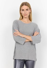 Femeie cu păr blond drept, îmbrăcată într-un pulover gri dintr-un material cu nervuri și pantaloni din piele negri, stand cu brațele încrucișate pe un fond alb.