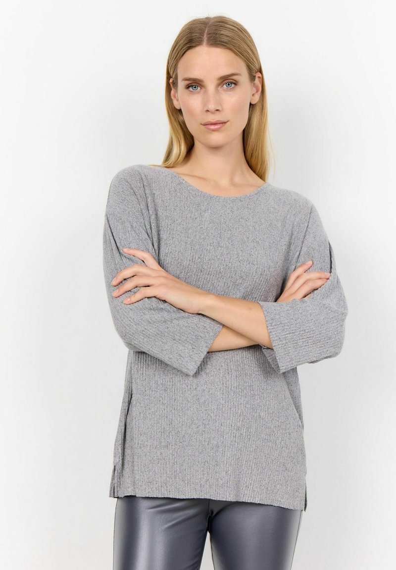 Femeie cu păr blond drept, îmbrăcată într-un pulover gri dintr-un material cu nervuri și pantaloni din piele negri, stand cu brațele încrucișate pe un fond alb.