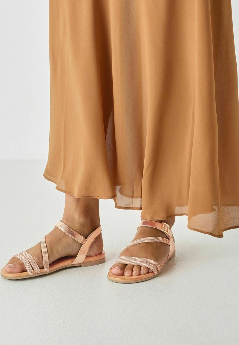 Sandales roses à brides avec une semelle plate, ornées de strass décoratifs et d'une boucle ajustable. Portées avec une robe fluide de couleur brun clair.