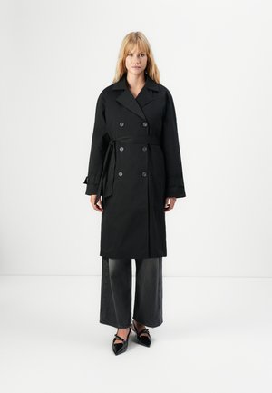 Woman wearing a black double-breasted trench coat with belt, wide-leg faded black jeans, and black pointed-toe heels.