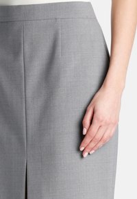 Grauer Bleistiftrock aus strukturiertem Stoff, mit tailliertem Bund, seitlichem Schlitz und scharfen Falten. Hand ruht leicht auf dem Stoff.