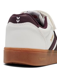 Witte sneaker met bruine zool, kastanjebruine hielkraag en Hummel-logo bij de zool en zijkant, gezien vanuit de achterzijhoek.