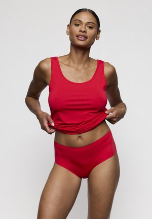 Femme aux cheveux foncés portant un débardeur rouge et une lingerie assortie, soulevant légèrement l'ourlet de son haut tout en regardant devant elle.