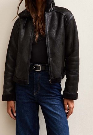 Donna che indossa una giacca di pelle nera con polsini e collo in shearling, top nero, jeans blu scuro e cintura nera con fibbia argentata.
