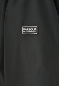 Μαύρο ύφασμα με απαλή υφή διαθέτει ορθογώνιο ετικέτα λογότυπου Barbour International σε λευκό και μαύρο.