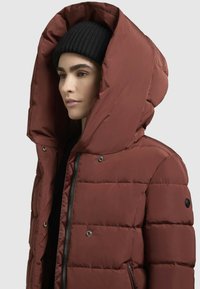 Frau, die eine große kapuzenförmige rostfarbene Steppjacke und eine schwarze Strickmütze trägt, die nach links gegen einen schlichten Hintergrund schaut.