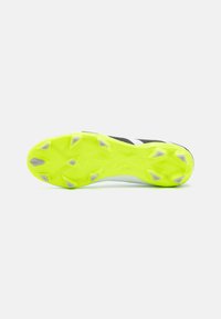 Chaussure de soccer noir et blanc avec une semelle jaune vif, dotée de pointes angulaires et d'une surface texturée pour une meilleure traction.