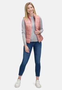 Jasnoróżowa pikowana kamizelka na szarym swetrze, w połączeniu z niebieskimi dżinsami i białymi trampkami. Kamizelka ma wysoki kołnierz i zamek błyskawiczny.