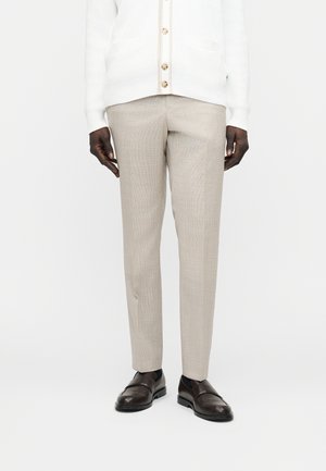 Mężczyzna w beżowych, dopasowanych spodniach, ciemnobrązowych formalnych skórzanych butach oraz białym, zapinanym na guziki kardiganie, stojący na białym tle.