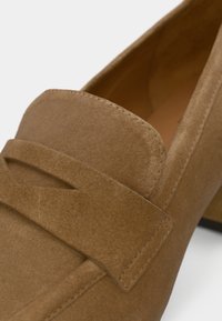 Καφέ μοκασίνια από καστόρι με ραμμένο σχέδιο και διακοσμητικό λουράκι στο πάνω μέρος. Η σόλα είναι ελαφρώς ανυψωμένη και λεία.