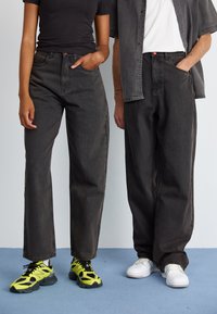 Mørke jeans på to figurer, hvor den ene har på en tætsiddende sort skjorte, mens den anden har en grå kortærmet skjorte på. Sorte og gule sko er synlige.
