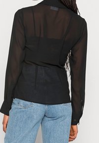Sort gennemsigtig bluse med lange ærmer, knapmanchetter og krave. Båret over blå jeans, der fremhæver et let, gennemskinneligt stof.