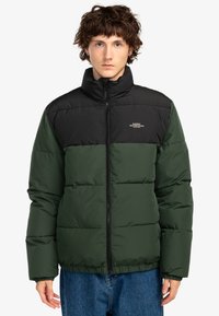 Ung person iført en sort og mørkegrøn puffjakke med frontlynlås og høj krave, parret med blå jeans, der står imod en hvid baggrund.