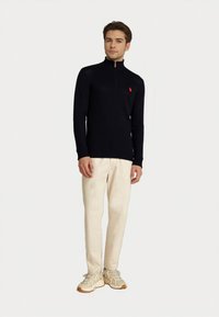 Mann steht und trägt einen schwarzen Reißverschluss-Pullover mit rotem Logo, beige Hosen und beige Sneakers vor einem schlichten weißen Hintergrund.