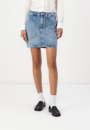 Kvinde iført en lyseblå denim mininederdel, hvid langærmet skjorte, hvide sokker og sorte loafers stående på en hvid gulv.