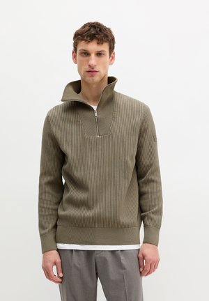 Mann trägt einen khakifarbenen, gerippten Reißverschluss-Pullover über einem weißen Unterhemd und grauen Hosen, steht vor einem schlichten weißen Hintergrund.