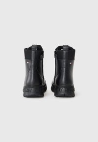 Bottines noires en cuir lisse avec des panneaux élastiques sur les côtés et une semelle texturée. À l'arrière, "TOMMY HILFIGER" est affiché en lettres grasses.