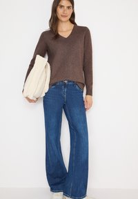 Brązowy sweter w serek, niebieskie szerokie jeansy, trzymając kremową kurtkę z polaru. Teksturowane materiały, luźny krój, jasne odcienie denimu.