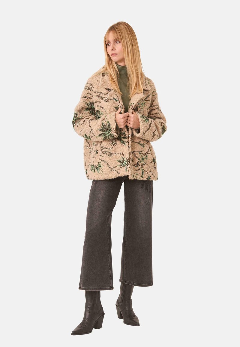 Chaqueta de piel beige con bordados botánicos verdes, diseño con cuello y botones delanteros, llevada con un suéter verde y pantalones de pierna ancha oscuros.
