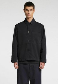 Mustast puuvillast jakk kraega, esikinnis nupuga ja ühe rinnataskuga. Kujundatud lõdva istuvuse ja pikkade varrukatega.