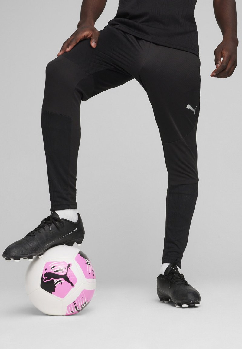 Zwarte sportbroek met textuurpanelen, gecombineerd met zwarte voetbalschoenen, staande op een witte en roze voetbal met een merlogo.