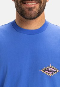 Blaues Baumwoll-T-Shirt mit einem kleinen mehrfarbigen Grafikdesign auf der linken Seite, das ein augenförmiges Motiv umgeben von stilisierten Strahlen zeigt.