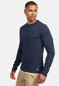 Marineblauwe geribbelde trui met lange mouwen en een ronde halslijn met een subtiel logo-accent; gedragen over een wit overhemd en gecombineerd met lichtgekleurde broek.