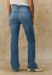 Vaqueros de campana en azul claro con un lavado medio, que presentan dos bolsillos traseros y una cintura clásica, combinados con zapatos beige de punta. 