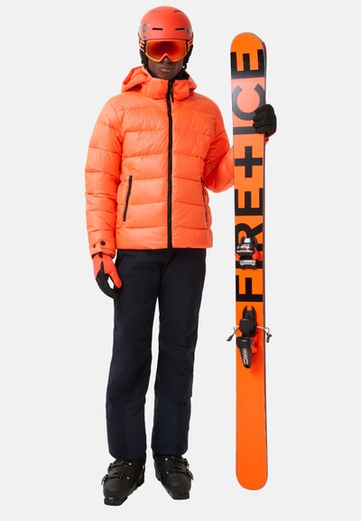 Jasnopomarańczowa kurtka puchowa i pasujący kask, czarne gogle narciarskie, granatowe spodnie narciarskie oraz czarne buty narciarskie trzymają pomarańczowo-czarną nartę.