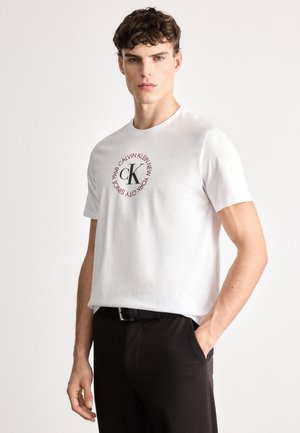 White cotton t-shirt featuring a circular logo with "CALVIN KLEIN NEW YORK" text and "1968" at the center, paired with dark pants.